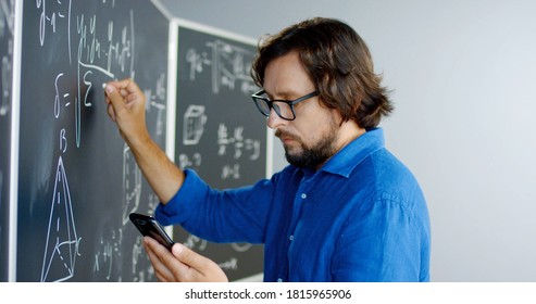 Close up of Caucasian male teacher in glasses writing formulas and math laws on blackboard and looking at smartphone. Educative mathematics lection. Man lecturer using mobile phone as cribe. - Powered by Shutterstock