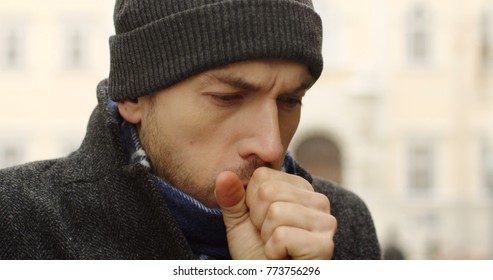 Close Up Of The Caucasian Man In A Black Hat Warming His Hands And Coughing Outside On The Winter Day. Blurred Background. Portrait.