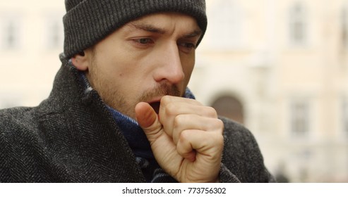 Close Up Of The Caucasian Man In A Black Hat Warming His Hands And Coughing Outside On The Winter Day. Blurred Background. Portrait.