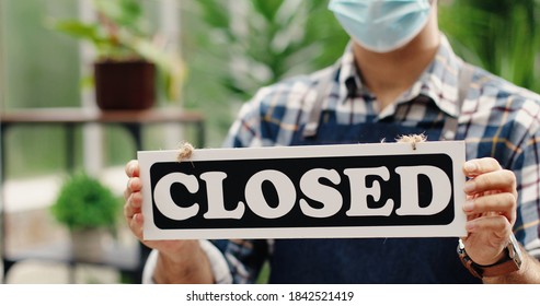 Close up of Caucasian man hands holding Closed sign indoors in florist shop in quarantine. Young male flower center employee in apron and mask standing in garden center with sign. Business concept - Shutterstock ID 1842521419