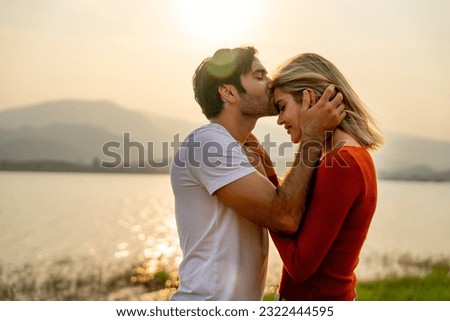 Close up Caucasian man kiss on 
forehead of beautiful woman with sunset light near lake and look romantic for couple love stay together.
