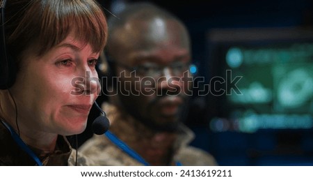 Close up of Caucasian middle-aged woman in headset talking via videochat in monitoring room in army. Female soldier having chat via webcam in military center. African American colleague on background.