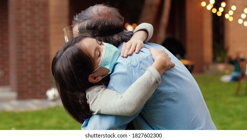 Close up of Caucasian small cute granddaughter in medical mask hugging her grandfather in yard. Barbeque meal on background. Little girl embracing grandpa in face shield outdoors. Pandemic concept.