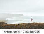 Close up caucasian woman in cold stand contemplates by the sea on viewpoint alone in windy moody day. Icelandic black sand beach atlantic ocean famous viewpoint