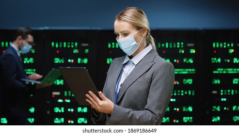 Close up of Caucasian woman security engineer in mask standing among servers with tablet device, tapping on screen and checking big data processors. Male analytic in data storage working on background