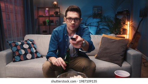 Close Up Of Caucasian Young Handsome Guy In Glasses Watching Television Choosing Channel While Sitting On Sofa And Drinking Beer From Bottle. Man Watch Movie On TV Sitting On Couch At Home, Leisure