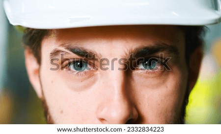 Close up of Caucasian young male engineer in white hardhat rising blue eyes and looking straight to camera. Factory worker. Indoors. Man in helmet. Manufactoring work concept. Industry concept.