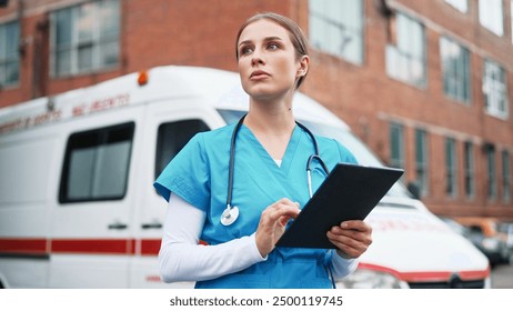 Close up of Caucasian young professional woman nurse texting on tablet browsing online standing outdoor near ambulance. Beautiful female doctor in blue uniform tapping on device and looking away