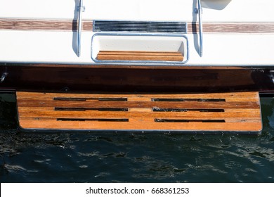 Close Up Centered Perspective Of Power Motor Water Skiing And Wake Boarding Fiberglass Boat Stern With Chrome Rail And Low Weathered Teak Wood Back Swim Step In Dark Blue Lake Water In Boating Scene