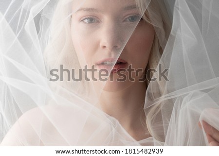 Close up of charming lady looking at camera and keeping lips slightly parted while covering face with tulle veiling