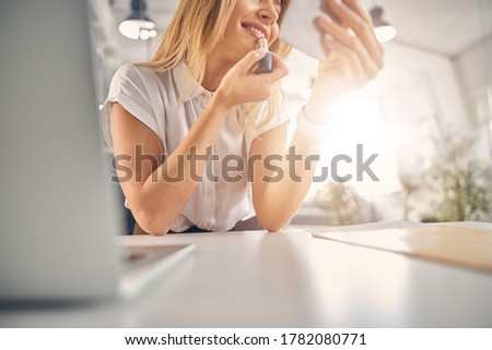Close up of charming lady sitting at the table and smiling while doing makeup in office