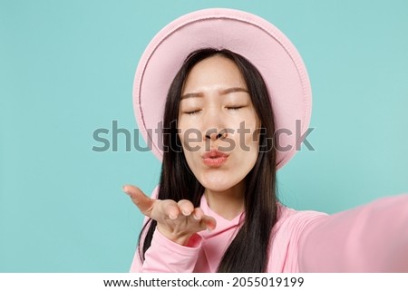 Close up charming young brunette asian woman 20s with eyes closed wears pink clothes hat doing selfie shot on mobile phone blow send air kiss isolated on pastel blue color background studio portrait.