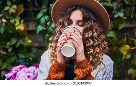 Close Up Of Charming Young Woman In Elegant Hat Enjoying Delicious Hot Drink On The Street. Stylish Woman With Curly Hair Drinking Coffee To Go While Spending Time Outdoors.