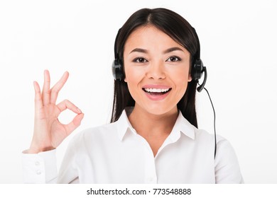 Close Up Of A Cheerful Asian Woman In White Shirt Wearing Headset With Microphone Showing Ok Gesture Isolated Over White Background