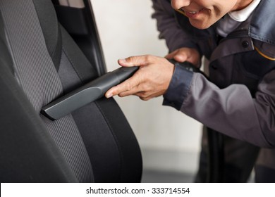 Close Up Of Cheerful Car Repair Shop Worker Is Cleaning The Interior Of The Car. He Is Holding A Vacuum Cleaner And Touching It To The Seat. The Man Is Smiling