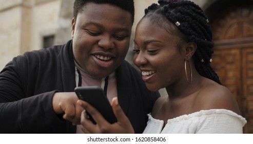 Close up of cheerful couple looking at smartphone screen and laughing. Young african couple standing outdoors using mobilephone, smiling, talking. Concept of people and coomunication