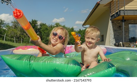 CLOSE UP Cheerful Little Children Floating In Pool And Spraying With Water Guns. Water Games For Hot Sunny Summer Days. Kids Enjoying And Having Fun While Playing In The Swimming Pool At Home Backyard