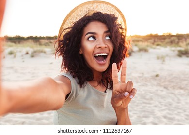 Close up of cheerful young african girl in summer hat taking a selfie at the beach and showing peace gesture - Powered by Shutterstock