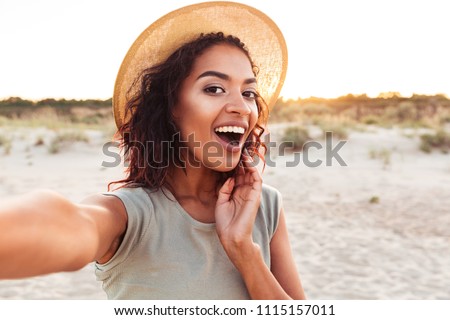 Close up of cheerful young african girl in summer hat taking a selfie at the beach