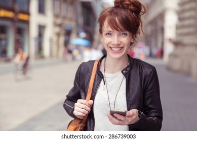 Close Up Cheerful Young Lady In Trendy Outfit, Holding Her Mobile Phone While Walking At The City Street And Looking At The Camera.