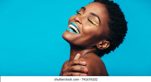 Close up of cheerful young woman with colorful makeup. Beauty portrait of female model with vivid makeup laughing on blue background.