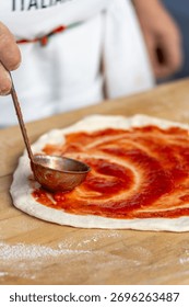 Close up of chef using a ladle to spread tomato sauce on pizza dough on a wooden board, capturing the authentic process of Italian food preparation.