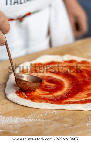 Close up of chef using a ladle to spread tomato sauce on pizza dough on a wooden board, capturing the authentic process of Italian food preparation.