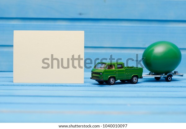 Close up of chicken egg on toy car with a trailer\
on a blue wooden background with blank card. Abstract retro\
concept. Selective\
focus\
