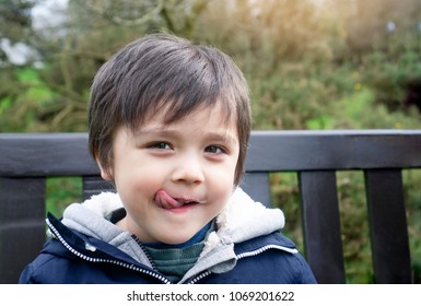 Close Up Child Face With Messy Mouth From Eating An Ice Cream, Cute Little Boy Sitting On Wooden Bench Looking At Camera With Licking His Lips, Head Shot Kid With A Dirty Face,Outdoor Activity Concept