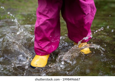 Close Up Of A Child Wearing Waterproof Boots And Trousers Splashing Through A Puddle Of Water.