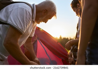 Close Up Of Children With Old Man Setting Up Red Tent Outside. Teacher With Gray Hair Hiking, Traveling, Organizing Campsite Together With Kids. Concept Of Active Rest.