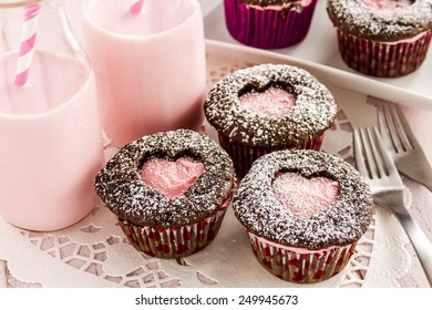 Close up of chocolate cupcakes with heart shaped cutout on top filled with pink frosting and two bottles of strawberry milk sitting on a white lace doily with two forks - Powered by Shutterstock