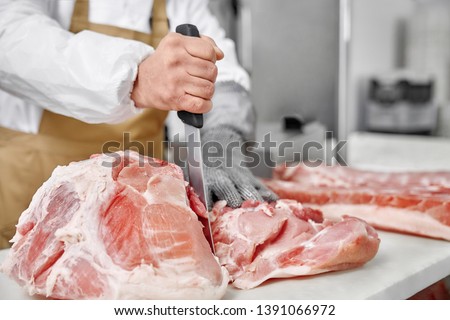 Close up of chopped pieces of meat and male hands of butcher in special gloves cutting fresh meat with knife. Meat pork or beef on table in butchery. Worker in white uniform and brown apron.