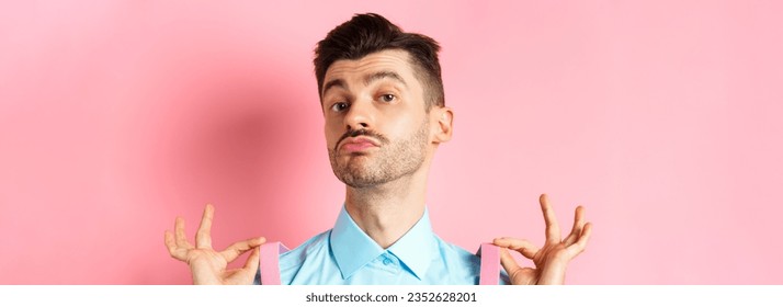 Close up of classy handsome guy adjusting suspenders, pucker lips and raise chin up, looking confident, standing in trendy outfit on pink background.