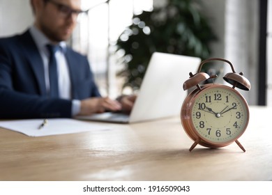 Close up of clock on forefront with businessman work on laptop online in office on background. Male employee prepare project at workplace, try meet deadline or appointment. Time management concept.