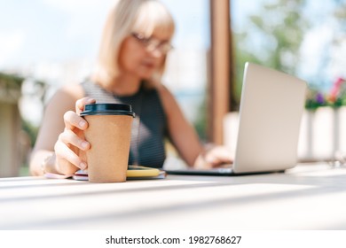 Close Up Coffe Cup. Adult Woman Typing At Laptop And Mobile Cell Phone Outside In Park. Thinking Senior Working And Drinking Coffee. Using Computer. Distance Learning Online Education And Shops.