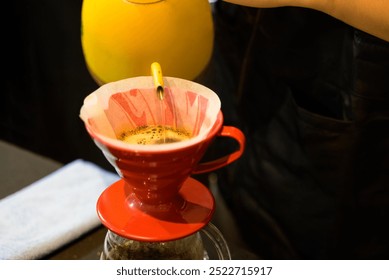 Close up of the coffee brewing process using drip pouring method. Hot water pouring from yellow kettle into red filter cone,with coffee grounds blooming inside paper filter
