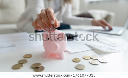 Close up coffee table lot of invoice, financial stats documents, utility bills, household payments, focus on female hand puts coin in piggy bank. Family budget manage, making personal savings concept