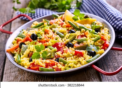 Close Up of Colorful and Fresh Vegetarian Paella Spanish Rice Dish Served in Pan with Red Handles and Linen Napkin on Rustic Wooden Table - Powered by Shutterstock