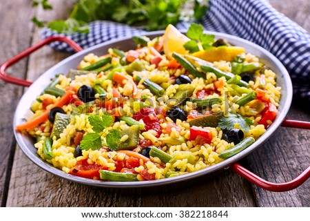Close Up of Colorful and Fresh Vegetarian Paella Spanish Rice Dish Served in Pan with Red Handles and Linen Napkin on Rustic Wooden Table