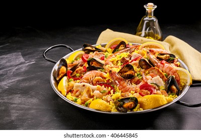 Close Up of Colorful Spanish Seafood Paella Rice Dish with Fresh Shellfish Served with Lemon Wedges in Pan on Smudged Chalkboard Background with Oil and Napkin