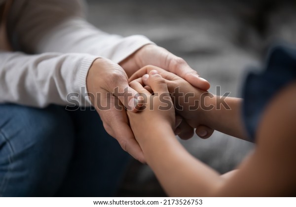Close up compassionate young foster parent holding\
hands of little kid girl, giving psychological help, supporting at\
home. Sincere different generations family sharing secrets or\
making peace.
