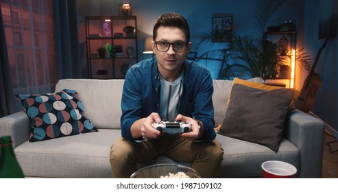 Close up of concentrated handsome young Caucasian guy in glasses sitting at home on sofa watching football game on TV and eating snacks. Man watch sport match on television, football fan concept