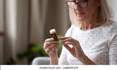 Close up concentrated middle aged woman wearing eyeglasses, holding bottle with pills in hands, reading instruction to medicine. Focused mature lady examining drug prescription label at home.