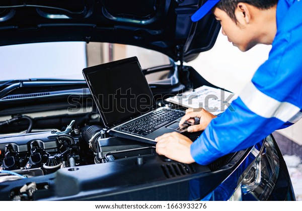 Close up concept of male asian car mechanic using a\
computer laptop to diagnosing and checking up on car engines parts\
for fixing and repair, working on the car engine wearing blue\
overall and a hat