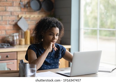 Close up confident African American woman looking at laptop screen, thoughtful student or businesswoman touching chin, pondering idea, strategy, working on research project in kitchen at home