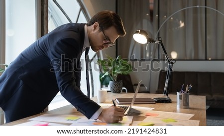 Close up confident businessman wearing glasses writing notes on colorful stickers, entrepreneur executive working with documents, analyzing financial project statistics, standing at table in office