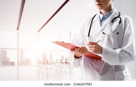 Close Up Of Confident Female Doctor In White Medical Uniform Making Notes Into Notebook While Standing Inside Of Bright Hospital Building. Medical Industry Concept