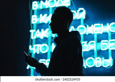 Close Up Of A Confident Young African Man Silhouette Wearing Windbreaker Using Mobile Phone While Standing Outdoors Close To A Neon Signboard