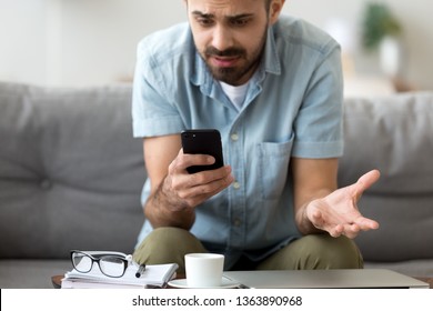 Close up confused man having problem with broken not working phone, annoyed frustrated male student receiving bad news, reading unpleasant email, sitting on sofa, looking at screen, spam message - Shutterstock ID 1363890968